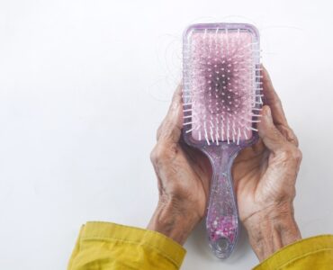 A rectangular hair brush with black hair in it on a white background. It is being cupped by the hands of a person wearing a yellow long sleeve shirt.